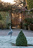 SEDGWICK PARK, WEST SUSSEX. ORNATE CAST IRON GATE (VISITORS ENTRANCE) TO GARDEN WITH FROSTED METAL BIRD SCULPTURE AND BOX TOPIARY SHAPE. WINTER, JANUARY, SUNLIGHT.