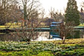 CHIPPENHAM PARK, CAMBRIDGESHIRE: SHEETS OF SNOWDROPS BESIDE THE LAKE WITH BOAT HOUSE IN BACKGROUND  -  WINTER
