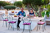 JONATHAN BAILLIE GARDEN, ALAIOR, MENORCA: JONATHAN AND JO BAILLIE AT THE OUTDOOR BUILT IN TABLE AND BARBEQUE WITH MENORCAN TERRIER DOTTIE. OUTDOOR LIVING, ENTERTAINING