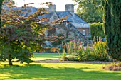 LAMPORT HALL, NORTHAMPTONSHIRE: VIEW TO THE ROCKERY ACROSS LAWN WITH BORDER OF DARK RED HOLLYHOCKS IN EVENING LIGHT. FORMAL, LAWN, CLASSIC COUNTRY HOUSE
