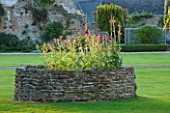 LAMPORT HALL, NORTHAMPTONSHIRE: WALLED RAISED BED - POSSIBLY A COCKPIT - PLANTED WITH CARDOONS AND HOLLYHOCKS - FLOWERS, FLOWERING. LAWN, CLASSIC ENGLISH GARDEN