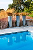 SURREY GARDEN DESIGNED BY ANTHONY PAUL: SWIMMING POOL WITH CAREX FLAGELLIFERA IN METAL CONTAINERS ON WOODEN PLINTHS. WATER, COUNTRY GARDEN, PATTERN, REFLECTION