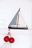 SALTWATER, NORFOLK : DESIGNER KAREN MOORE - CHRISTMAS, DECEMBER, WINTER - BEDROOM IN RED AND WHITE - TOY WOODEN BOAT ORNAMENT ON WALL WITH RED BAUBLES - ORNAMENT, DECORATION