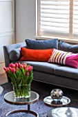 SALLY STOREY HOUSE, LONDON: OPEN PLAN SITTING ROOM / HALL WITH GREY SETTEE, RUG AND GLASS TABLE WITH TULIPS IN VASE, CUSHIONS, SHUTTERS