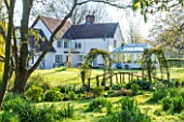 ULTING WICK, ESSEX: VIEW ACROSS LAWN TO THE HOUSE WITH WOODEN BRIDGE IN SPRING. APRIL