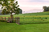 GLYNDEBOURNE, EAST SUSSEX: VIEW PAST WOODEN FENCE AND WOODEN BENCH TO THE COUNTRYSIDE BEYOND AT DAWN. SUMMER, MIST