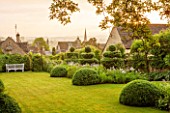 GREYHOUNDS, BURFORD, OXFORDSHIRE:DAWN LIGHT ON CLASSIC COUNTRY GARDEN WITH LAWN, BOX DOMES AND YEW TOPIARY. BORDERS WITH COTTAGE GARDEN PLANTS. WOODEN BENCH. A PLACE TO SIT. SUMMER