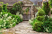 OXLEAZE FARM, OXFORDSHIRE: STONE PAVED PATH LEADING TO OLD WOODEN & RUSTED METAL ORNATE GARDEN GATE WITH CENTRANTHUS RUBER ALBUS (WHITE VALERIAN) & VARIGATED BOX TOPIARY SHAPES