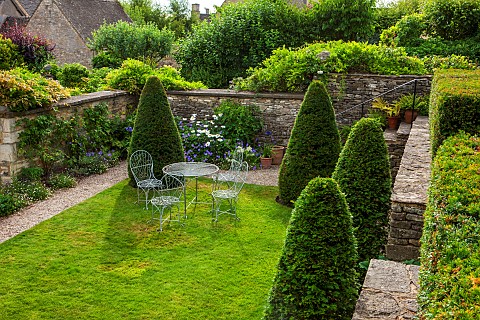 CALENDARSBURFORDOXFORDSHIRECLIPPED_YEW_CONES_ON_LAWN_WALL_WITH_TABLE__CHAIRSBORDER_WITH_ERIGERON_KAR