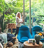 ABIGAIL AHERN HOUSE, LONDON: ABIGAIL AHERN IN THE LIVING ROOM - BLUE CHAIRS, NILE OTTOMAN STOOL, FAKE GOLDENBALL CACTUS, PATIO OUTSIDE, INSIDE OUT, SETTEE, MAUD THE DOG, PET