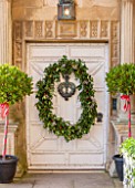 BURTON AGNES HALL, EAST YORKSHIRE: CHRISTMAS - WHITE FRONT DOORWAY WITH BAY TREES IN BLACK CONTAINERS, HOLLY AND BERRY WREATH MADE BY THE GARDENERS WITH PLANTS FROM ESTATE