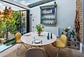LONDON HOUSE DESIGNED BY JULIE SIMONSEN. MORNING ROOM WITH GLASS BIFOLD DOORS  LEADING TO GARDEN. CIRCULAR TABLE LAID FOR BREAKFAST WITH CONTEMPORARY YELLOW CHAIRS. DISTRESSED WALL SHELF AND COLOURED GLASSWARE