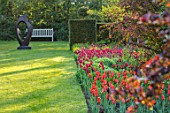PASHLEY MANOR GARDEN, EAST SUSSEX. SPRING, BORDER, TULIPS - TULIPA BALLERINA, TULIPA ILE DE FRANCE. BULBS, COUNTRY, ORANGE, RED, FLOWERS, FAMILY CIRCLE SCULPTURE BY JOHN BROWN