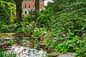 MORTON HALL, WORCESTERSHIRE: THE UPPER POOL IN THE STROLL GARDEN WITH HOUSE BEHIND. SPRING, POND, WATER, SHADE, SHADY, WOODLAND, PRIMULA PULVERULENTA