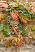 THE CONIFERS, OXFORDSHIRE: DESIGNER CLIVE NICHOLS - HEAD BY FIONA BARRATT ON WALL WITH VITIS COIGNETIAE. COTSWOLDS, ORNAMENT, COTTAGE, AUTUMN, FALL, OCTOBER, SHRUBS, FOLIAGE