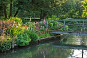 ABLINGTON MANOR GLOUCESTERSHIRE: VIEW ACROSS COLN RIVER TO ISLAND BED AND WOODEN FOOTBRIDGE. REFLECTIONS WATER. CLASSIC COUNTRY GARDEN COTSWOLDS ROMANTIC ROMANCE LAWN