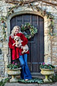 PYTTS HOUSE, OXFORDSHIRE: CHRISTMAS, BACK DOOR, WREATH, ANNA DE KEYSER HOLDING ONE OF HER PET DOGS