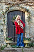 PYTTS HOUSE, OXFORDSHIRE: CHRISTMAS, BACK DOOR, WREATH, ANNA DE KEYSER HOLDING ONE OF HER PET DOGS