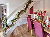 THE CONIFERS, OXFORDSHIRE: CHRISTMAS - KITCHEN DINING ROOM - TABLE, CHAIRS, PRINT OF RED CAMELLIA BY CLIVE NICHOLS, STAIRCASE, VASE WITH AMARYLLIS, PINEAPPLE CANDLES
