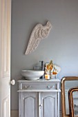 JACKY HOBBS HOUSE, LONDON: BATHROOM IN GREY. GREY VINTAGE WOODEN WASHSTAND, CERAMIC BOWL, CARVED PLASTER ANGELS WING ON WALL. CANDLE