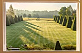 ROCKCLIFFE GARDEN, GLOUCESTERSHIRE: VIEW ACROSS LAWN AT SUNRISE WITH TERRACE, CLIPPED BEECH OBELISKS, BORROWED LANDSCAPE, BRONZE SCULPTURE SOUTHERN SHADE BY NIGEL HALL