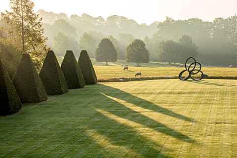 ROCKCLIFFE_GARDEN_GLOUCESTERSHIRE_VIEW_ACROSS_LAWN_AT_SUNRISE_WITH_TERRACE_CLIPPED_BEECH_OBELISKS_BO