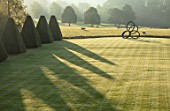 ROCKCLIFFE GARDEN, GLOUCESTERSHIRE: VIEW ACROSS LAWN AT SUNRISE, CLIPPED BEECH OBELISKS, BORROWED LANDSCAPE, BRONZE SCULPTURE SOUTHERN SHADE BY NIGEL HALL, SHADOWS