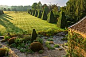 ROCKCLIFFE GARDEN, GLOUCESTERSHIRE: VIEW ACROSS LAWN AT SUNRISE WITH TERRACE, CLIPPED BEECH OBELISKS, BORROWED LANDSCAPE, BRONZE SCULPTURE SOUTHERN SHADE BY NIGEL HALL
