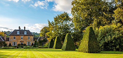 ROCKCLIFFE_GARDEN_GLOUCESTERSHIRE_VIEW_ACROSS_LAWN_TO_HOUSE_CLIPPED_BEECH_OBELISKS_ENGLISH_COUNTRY_G