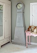 PEAR TREE COTTAGE, OXFORDSHIRE: MASTER BEDROOM, BLUE, GREY VINTAGE SWEDISH MORA CLOCK, PAINTED ARMOIRE, DISTRESSED PINK SIDE TABLE WITH COLLECTION OF VINTAGE TOYS