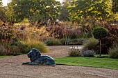 GRANTLEY HALL, YORKSHIRE: GRAVEL GARDEN, CURVED WOODEN SEAT, BENCH, A PLACE TO SIT, STIPA GIGANTEA, WOODS, TREES, LION STATUE, LAWN