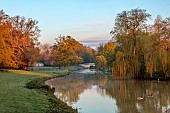 COTTESBROOKE HALL AND GARDENS, NORTHAMPTONSHIRE: SPRING, APRIL, VIEW OF PARKLAND, LAKE AND BRIDGE FROM THE MITCHELL BRIDGE, SUNRISE