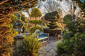 THE LASKETT GARDEN, HEREFORDSHIRE: WINTER, FROST, JANUARY, VIEW THROUGH YEW HEDGE TO WILD GARDEN, STONE URN ON PLINTH, BLUE WOODEN BENCH, SEAT, CLIPPED YEW TOPIARY, PATH