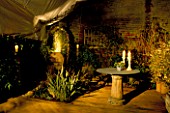 CERAMIC CHAIR & CERAMIC/GLASS TABLE IN NIGHT LIT CORNER OF GARDEN WITH CANDLES & UPLIGHTING. DESIGNER: EMMA LUSH. WATER FEATURE BY MARK LAURENCE