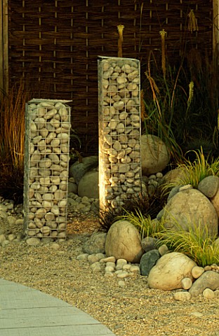 SIMPLE_SCULPTURES_OF_TALL_WIRE_COLUMNS_FILLED_WITH_STONES_STAND_AMONGST_GRASSES_AND_GRAVEL_IN_MODERN