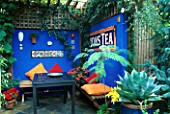 COURTYARD PART COVERED BY A TIMBER PERGOLA. TABLE AND BENCHES WITH ORANGE AND RED CUSHIONS BESIDE ULTRAMARINE WALLS DECORATED WITH MOSAIC PANELS AND MEXICAN TILES. KARLA NEWELL
