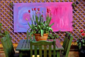 A PLACE TO SIT : GREEN TABLE & CHAIRS WITH TERRACOTTA CONTAINER PLANTED WITH RED TULIPS . IN THE B/GROUND IS ABSTRACT ART ON PURPLE TRELLIS. THE NICHOLS GDN  READING