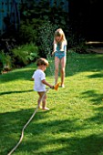 HAZEL AND WILLIAM PLAYING WITH A HOSEPIPE IN THE GARDEN. THE NICHOLS GARDEN  READING