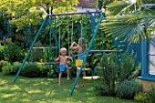 HAZEL AND ROBERT PLAYING ON THE SWING WITH THE BLUE FENCE AND GOLDEN HOP BEHIND. THE NICHOLS GARDEN  READING