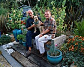 ROBIN GREEN AND RALPH CADE WITH THEIR DOG RUFUS SIT ON THE WEATHERED WOODEN HERB BENCH IN THEIR SEASIDE STYLE GARDEN  LONDON.