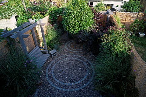 OVERVIEW_OF_SMALL_GARDEN_WITH_CIRCULAR_BRICK_PATTENS_IN_GRAVEL_NATURAL_WOODEN_SWING_SEAT_AND_STOOL__