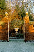 VIEW THROUGH THE GATES TOWARDS AN ORNAMENTAL URN LIT BY WINTER SUNLIGHT. THE FROST COVERED GARDEN AT WOLLERTON OLD HALL  SHROPSHIRE.