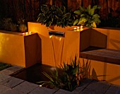 GARDEN LIGHTING:  WATER FEATURE: POST BOX STYLE FOUNTAIN SURROUNDED BY YELLOW RENDERED WALLS WITH BENCH SEAT AND HOSTAS IN BACKGROUND  AND NIGHT LIGHT CANDLES.  DESIGNER JOE SWIFT.