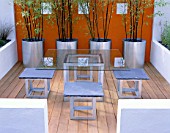 MINIMALIST GARDEN BY WYNNIATT-HUSEY CLARKE: GLASS TABLE WITH METAL AND SLATE CHAIRS ON WOODEN DECKING WITH GALVANISED  METAL CONTAINERS PLANTED WITH BLACK STEMMED BAMBOO