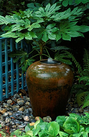 WATER_FEATURE_TERRACOTTA_POT_SURROUNDED_BY_PEBBLES_WITH_FATSIA_JAPONICA_BEHIND_DESIGNER_SARAH_LAYTON