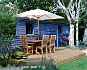BLUE SUMMERHOUSE WITH DECKING  TABLE  CHAIRS AND PARASOL  BIRCH TREE TRUNKS  ORNAGE LILLIES  HAMMOCK AND GRAVEL. DESIGNER: CLARE MATTHEWS