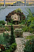 VIEW ALONG GARDEN PAST BOX AND TRACHYCARPUS TO WATER FEATURE MADE OF BRICK WALL AND  BRIGHTON PIER GOD HEAD WITH SHELLS BACKED BY ROCKS AND BLUE WALL WITH TRELLIS. DESIGNER ANDREW