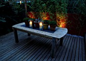DECKED TERRACE WITH LIGHTING: WOODEN TABLE WITH CANDLES AND TERRACOTTA POTS WITH RED WINE COLOURED WALL AND PHYLLOSTACHYS AUREA. LIGHTING BY GARDEN & SECURITY LIGHTING