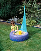 JOSHUA PLAYS IN THE TYRE SANDPIT ON THE LAWN. THE SAIL IS MADE OF KITE MATERIAL