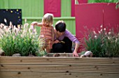 CHILDRENS DECK GARDEN: LUCY AND RACHAEL PLAY IN THE RAISED WOODEN BED. THE GRASS IS PENNISETUM HAMELYN